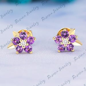 May include: A pair of gold flower stud earrings. Each earring features a cluster of purple gemstones arranged in a floral pattern, with a small white gemstone at the center. The earrings have a gold leaf design.