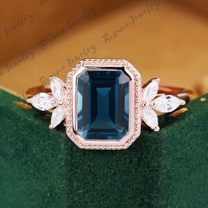 May include: A rose gold engagement ring with a large, emerald-cut blue topaz gemstone surrounded by a delicate halo of smaller diamonds. The ring is set on a white band and features a floral design on either side of the center stone.