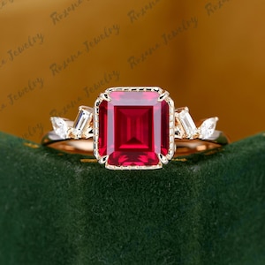 以下が含まれることがあります： 正方形のルビーがセットされたゴールドのバンドの婚約指輪。側面には小さなダイヤモンドが並んでいます。指輪は緑色のベルベットの箱の上に置かれています。