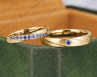 May include: Two gold rings with a polished finish. One ring features a row of small, round blue stones set along the band. The second ring has a single, round blue stone set in the center of the band. Both bands have a smooth, curved design.