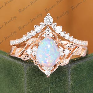 以下が含まれることがあります： ローズゴールドの婚約指輪セット。洋梨型のオパールをセンターに、ダイヤモンドのハローとダイヤモンドをちりばめたバンドで囲んでいます。セットには、ダイヤモンド付きの王冠型のバンドが含まれています。リングはフローラルデザインです。