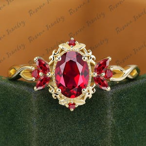 May include: A gold ring featuring an oval-cut, deep red gemstone at the center, surrounded by smaller red gemstones and ornate gold detailing. The ring's band has a twisted design. The words "Roxana Jewelry" are in the background.