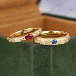 May include: Two gold bands featuring intricate engraved scrollwork patterns. One ring is set with a single marquise-cut red gemstone, while the other ring features a single round-cut blue gemstone. Both rings have a polished gold finish.