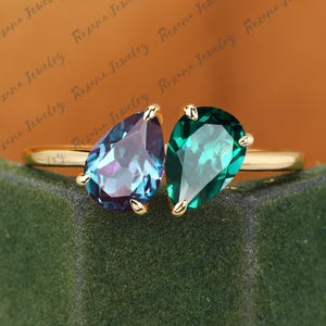 Könnte beinhalten: Ein goldener Ring mit zwei tropfenförmigen Edelsteinen. Einer ist tief türkis, der andere leuchtend smaragdgrün. Der Ring liegt auf einem grünen Samthintergrund, mit dem Schriftzug "Roxana Jewelry".