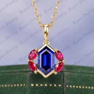 May include: A gold pendant necklace featuring a hexagonal blue gemstone center, surrounded by red gemstones. The necklace has a delicate gold chain. The Roxana Jewelry logo is visible in the background.