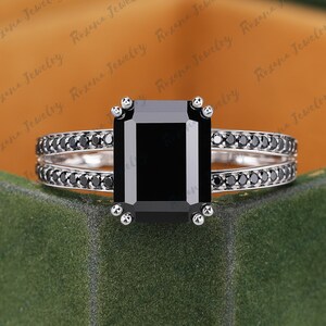 May include: A silver-toned ring featuring a large, rectangular, emerald-cut black center stone held by four prongs. The split shank band is adorned with a row of small, round, black accent stones along each side of the metal band structure.