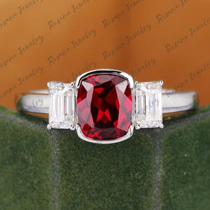 Puede incluir: Un anillo de plata con una gran piedra preciosa roja oscura de talla cojín, flanqueada por dos piedras preciosas rectangulares y transparentes. El anillo está sobre una superficie de terciopelo verde. El texto "Roxana Jewelry" es visible en el fondo.