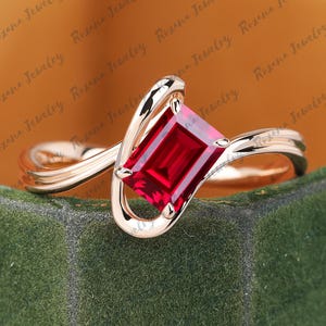 Puede incluir: Un anillo de oro rosa con una gema rectangular de color rojo intenso. La banda del anillo tiene un diseño único y ondulado que acuna la gema. El anillo se exhibe sobre una superficie de terciopelo verde, con las palabras "Roxana Jewelry" en el fondo.