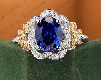 Op de afbeelding: Een blauwe saffier verlovingsring met een bloemmotief. De ring is gezet in witgoud en heeft een halo van diamanten rond de middelste steen. De band is geaccentueerd met geelgoud en kleine diamanten.