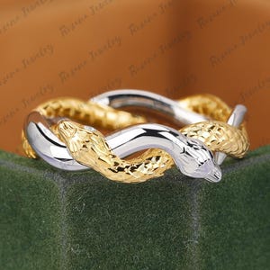 May include: A ring featuring two intertwined snakes, one in a polished silver-tone metal and the other in a textured gold-tone metal. The snake heads are positioned at the front, with detailed scale patterns visible on the gold-tone body.