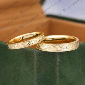 May include: Two gold wedding bands with a floral engraved design. The bands are on a green velvet surface.