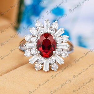 May include: A silver ring featuring a large, oval-cut, deep red gemstone at the center, surrounded by a sunburst design of white baguette and round-cut stones. The ring has a split shank design and is set against a neutral background.