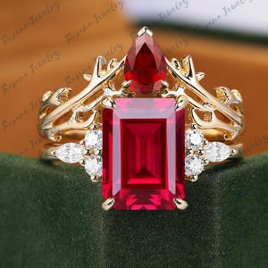 May include: A gold ring with a large, rectangular ruby center stone. The ring features a crown-like design with a pear-shaped ruby and small white stones. The ring is set against a green background.