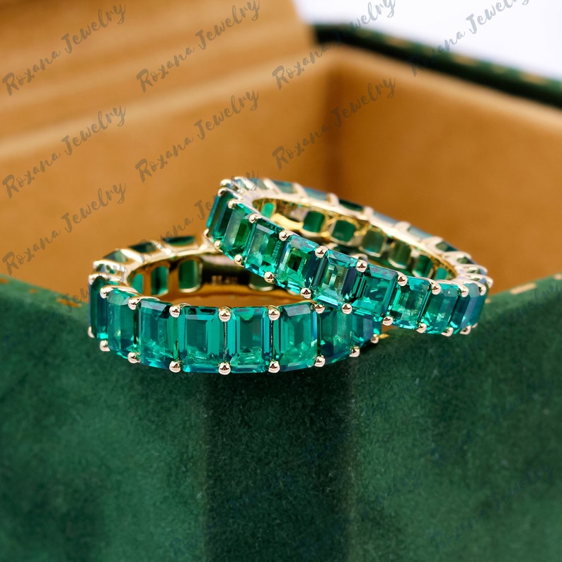 May include: Two emerald green eternity rings with square-shaped stones set in a gold band.