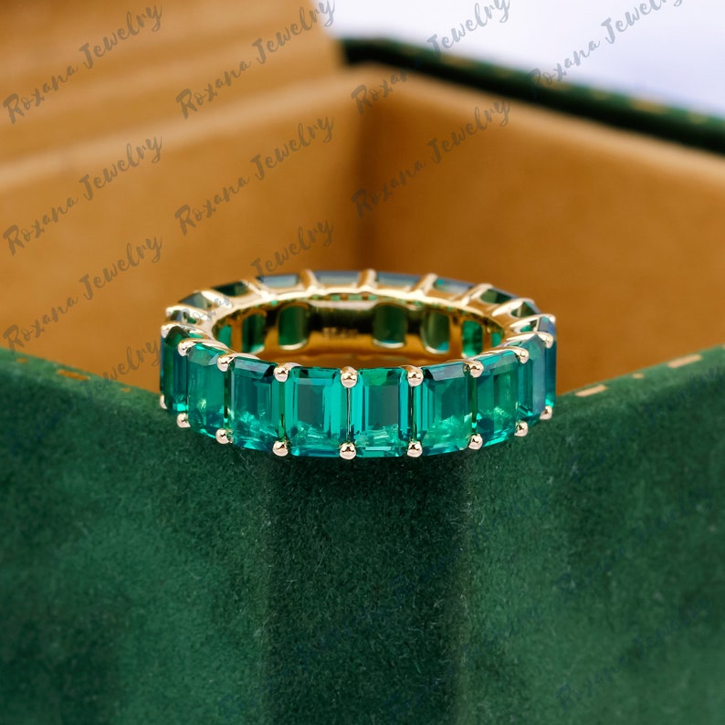 May include: A gold band ring with a row of emerald green rectangular gemstones. The ring is set on a green velvet surface.