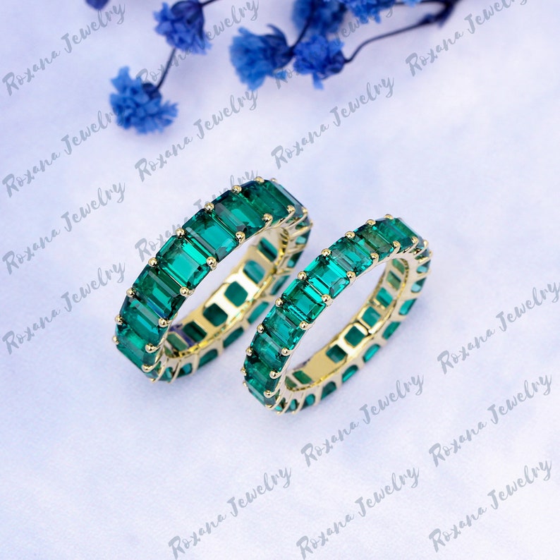 May include: Two emerald green eternity bands with square-shaped stones set in a yellow gold band.