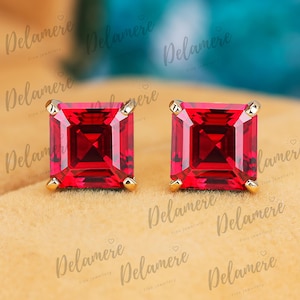 May include: A pair of square-cut, vibrant red gemstone stud earrings with gold settings. The earrings are displayed on a light tan surface, with the Delamere Fine Jewellery logo subtly visible in the background.