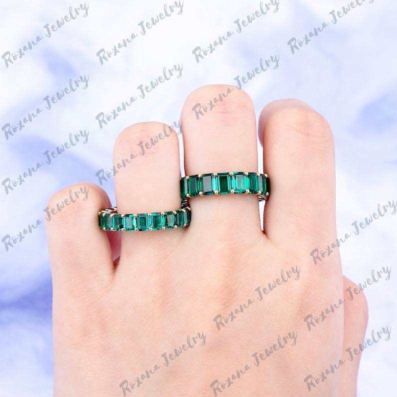 May include: Two emerald green eternity rings with square-shaped stones set in a gold band. The rings are worn on the index and middle fingers of a hand.