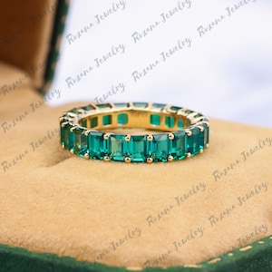 May include: A gold band ring with a row of emerald green square-shaped gemstones.