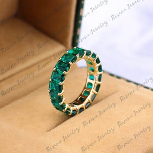 May include: A gold band ring with emerald green square-shaped gemstones set in a continuous line around the band.