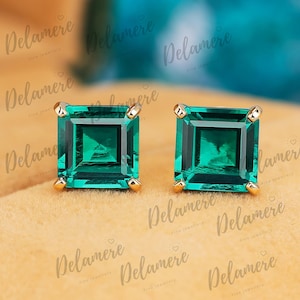 May include: A pair of square-cut emerald stud earrings with gold settings. The emeralds are a vibrant green color and are set in a four-prong gold setting. The earrings are displayed on a tan surface.