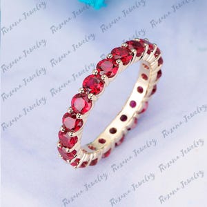 May include: A gold band ring with a row of red gemstones. The ring is set with round, red gemstones that are evenly spaced around the band.