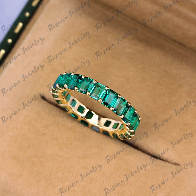 May include: A gold band ring with a row of emerald green square-shaped gemstones.