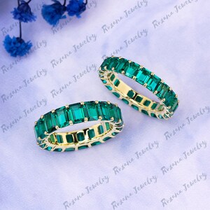May include: Two gold eternity bands with emerald green square-shaped gemstones. The bands are set with a continuous row of stones.