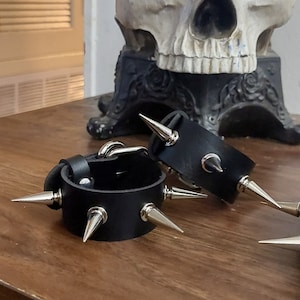 May include: Two black leather wrist cuffs with silver metal spikes. The cuffs are on a wooden table in front of a white skull with a red flower crown.
