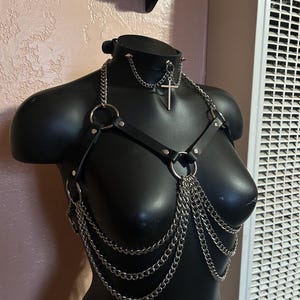 May include: A black leather harness with silver chains and a silver cross pendant. The harness has two rings on the front and a choker with a silver cross pendant.