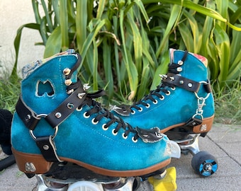 May include: A pair of teal blue roller skates with black leather straps and silver studs. The skates have black laces and white wheels.