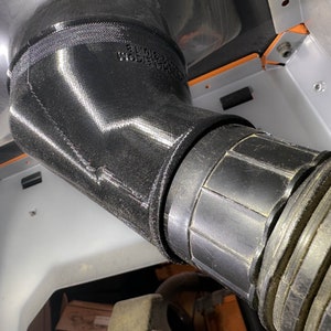 May include: Close-up of a black rubber hose connected to a black plastic pipe. The pipe has a ribbed texture and is connected to a larger, black, 3D printed pipe.