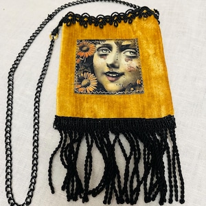 May include: Mustard yellow velvet pouch with a black chain strap and fringe. The pouch features a printed image of a woman's face surrounded by flowers. The top edge has a black lace trim.