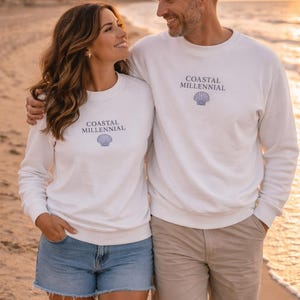 May include: Two people wearing white sweatshirts with the text "COASTAL MILLENNIAL" and a seashell graphic. The woman wears denim shorts, and the man wears khaki shorts. They are walking on a beach at sunset.