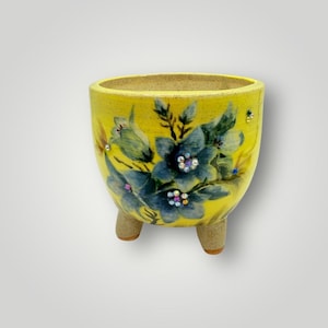 May include: A yellow ceramic planter with three small feet. The planter is decorated with blue floral designs and small, colorful embellishments. The interior and feet have a natural, unglazed finish.