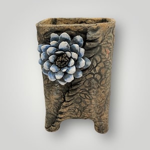 May include: A handcrafted, textured ceramic vase with a square top and two small feet. The vase is adorned with a blue and white flower on the front. The vase has a brown and gray color scheme and a unique, artistic design.