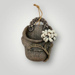 May include: A handcrafted, gray ceramic wall planter with a braided handle and a jute rope for hanging. The planter features a sculpted white flower and leaf design, adding a touch of nature-inspired decor. Ideal for small plants or succulents.