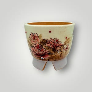 May include: A cream-colored ceramic planter with a floral design featuring pink roses and brown leaves. The planter has a light brown rim and is adorned with small red dots and rhinestones. It has a unique, curved base.