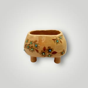 May include: A small, beige ceramic planter with three short legs. The planter is decorated with brown floral designs and teal-colored embellishments. The open top is rectangular, and the overall shape is rounded.