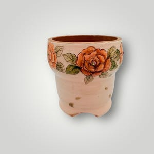 May include: A handcrafted, peach-colored ceramic planter with a decorative floral design. The planter features a series of orange roses with green leaves around the rim. The planter has three small feet and several small holes.