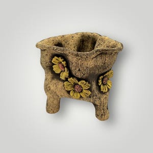 May include: A textured, tan-colored planter with three small legs and a wavy rim. The planter is adorned with three yellow and pink flower appliqués. The overall design gives the appearance of a rustic, handmade item, suitable for small plants or succulents.