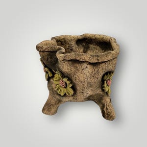 May include: A decorative, three-legged planter with a textured, earthy brown finish. The planter features embossed yellow flowers with pink centers. The planter has a unique, organic shape, perfect for displaying small plants or flowers.
