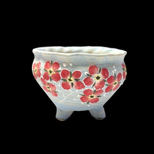 May include: A light blue ceramic planter with three small feet. The planter is decorated with red flowers with yellow centers. White dots are painted around the flowers. The rim of the planter has a wavy edge.