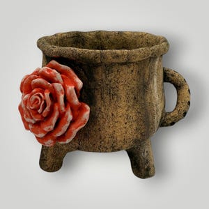 May include: A brown ceramic planter with a textured finish, featuring a vibrant red rose applique. The planter has three small legs and a handle, adding a touch of rustic charm. Ideal for small plants or succulents.