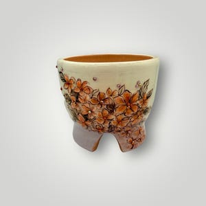 May include: A cream-colored ceramic planter with a floral design in shades of orange and brown. The planter has three small feet and is adorned with small, sparkling embellishments. The interior is a warm, earthy tone.