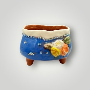 May include: A handcrafted blue ceramic planter with a white rim and three small feet. The planter is decorated with colorful floral designs and sparkling embellishments. A unique piece for home decor.
