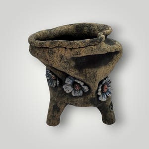 May include: A rustic, textured, brown and tan ceramic planter with a unique, asymmetrical shape. The planter features three-dimensional, light blue and pink floral accents on its legs. The open top suggests it is designed for plants or decorative items.