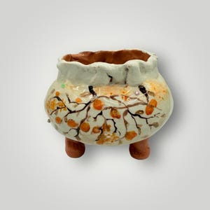 May include: A handcrafted ceramic pot with a cream-colored exterior, adorned with orange fruit and brown branch designs. The pot has a ruffled rim and three brown feet. The interior is a warm brown color.