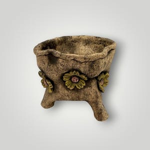 May include: A decorative, three-legged planter with a textured, earthy brown finish. The planter features three embossed flower designs with green petals and pink centers. The rim has a wavy edge, adding to its rustic charm.
