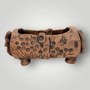 May include: A rectangular terracotta planter with a textured surface. The planter features a floral and butterfly design. The planter has two handles and rests on four small feet. The planter is approximately 10 inches long.